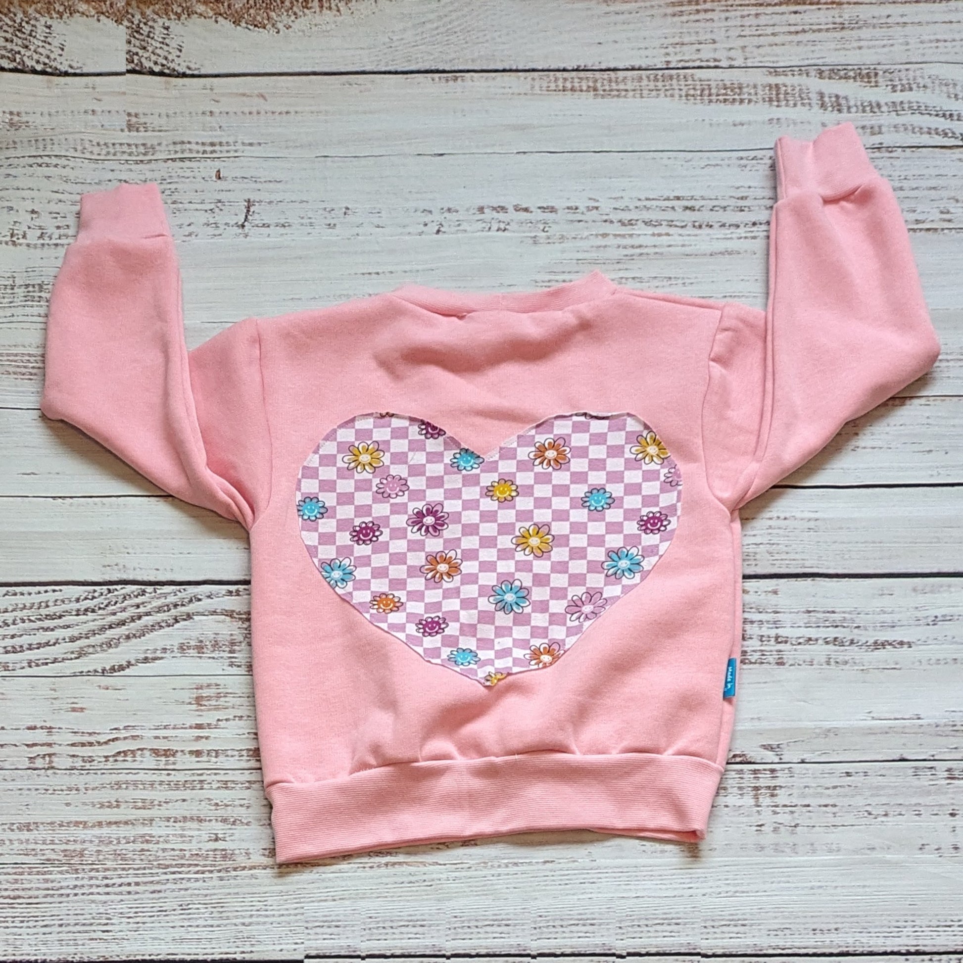 Pink checked happy flowers heart sweatshirt. Handmade in your colour choice of cotton French terry, with matching cotton ribbing and fun pink checked smiling flowers heart detailing on the back. Shown in pink French terry from the rear.