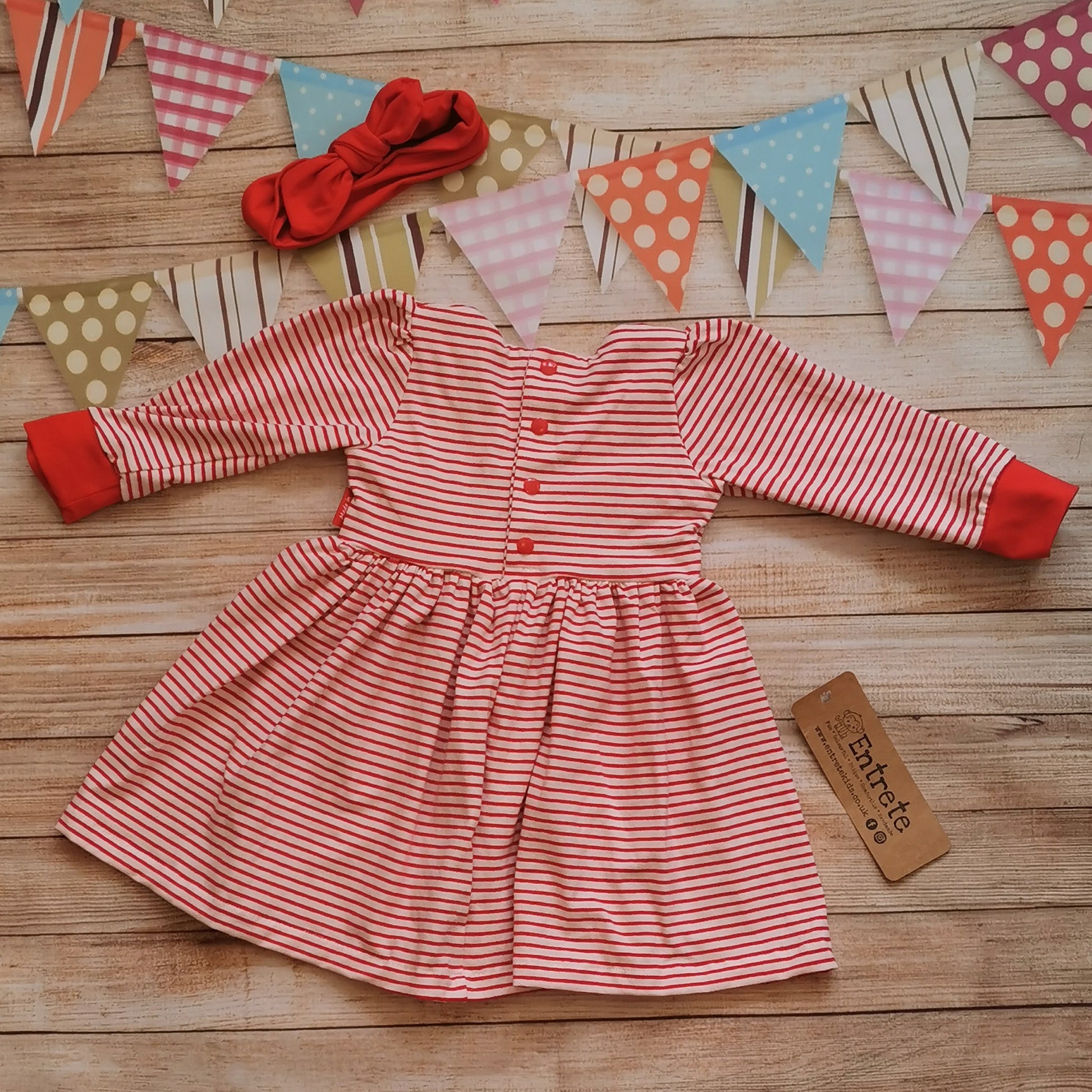 Rear view of the pretty red bow dress. Handmade using red striped cotton jersey and red cotton jersey, with adorable red bow detailing to the front. Shown with a matching red headband (sold separately)