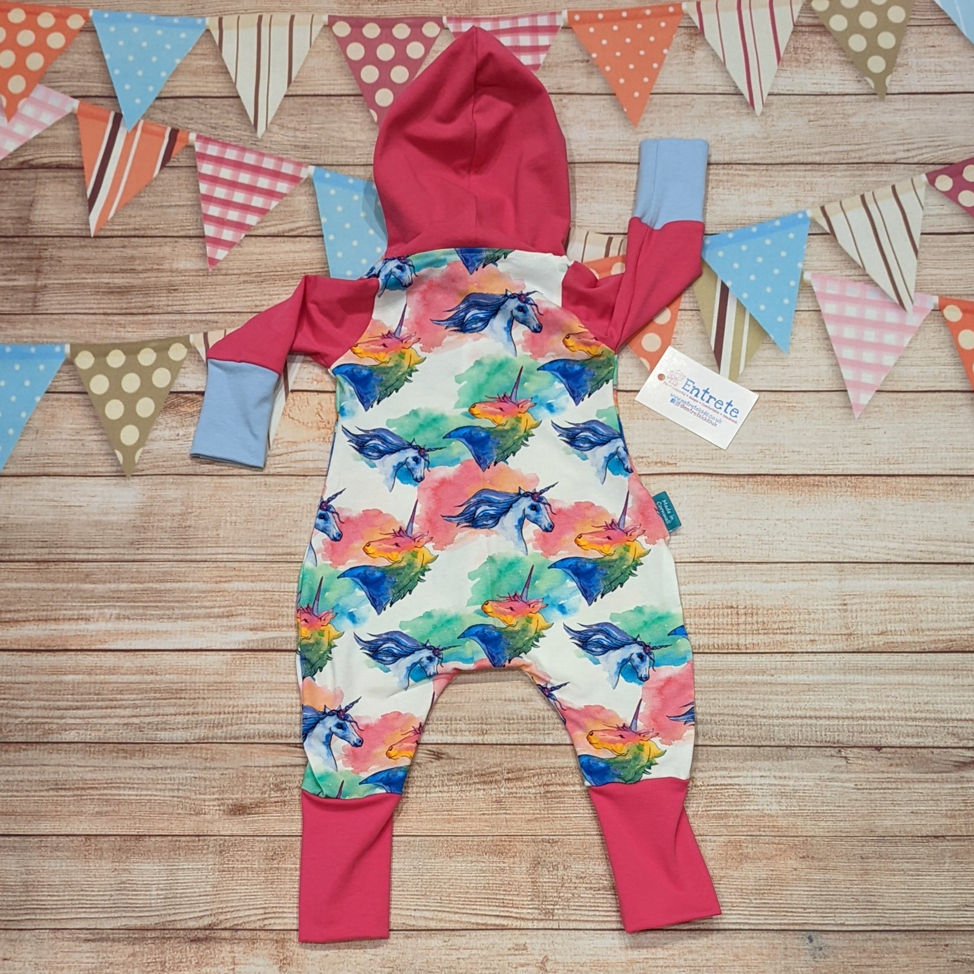 The colourful unicorns on white hooded romper. Handmade using colourful unicorns on white, fuchsia and sky blue cotton jerseys' with cream cotton ribbing. Shown from the rear.