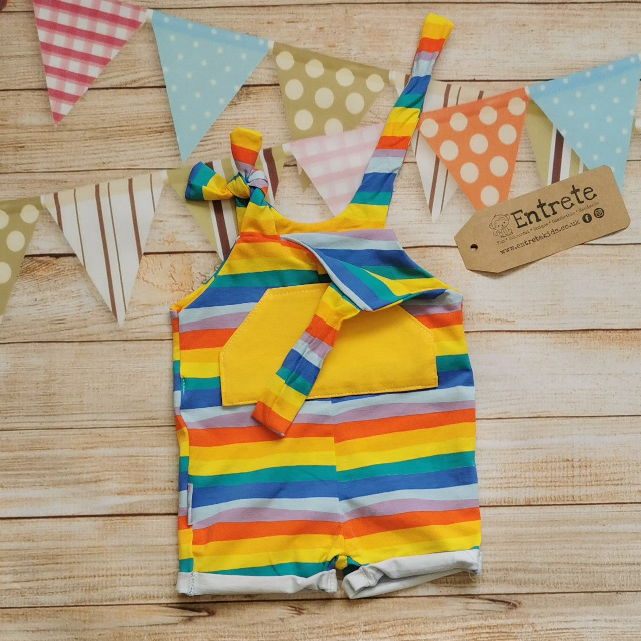 Unisex Kids Tie Strap Dungaree Shorts, handmade using red rainbow striped cotton jersey and yellow organic cotton jersey. With rolled cuffs and tie shoulder straps that are infinitely adjustable, perfect for growing kids. Showing an open strap.